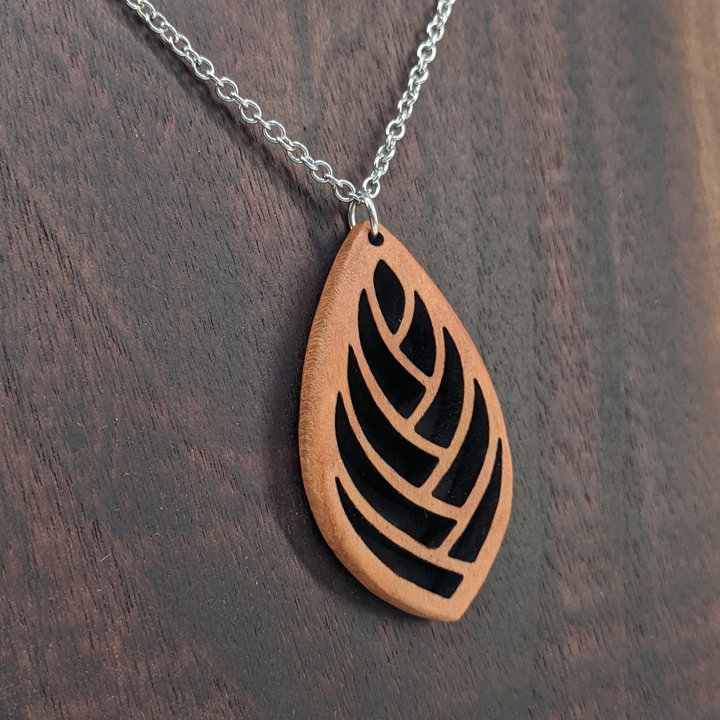 Wooden necklace pendant cut into a teardrop shape with a herring bone pattern cut throughout it. Made from cherry and hanging from a silver stainless steel chain against a dark walnut background.