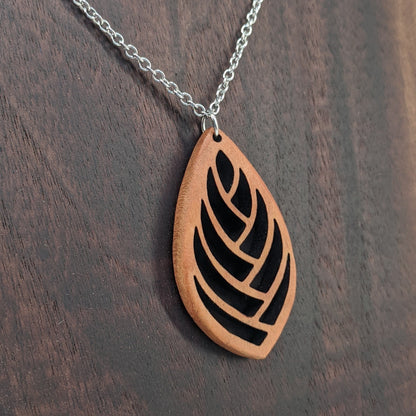 Wooden necklace pendant cut into a teardrop shape with a herring bone pattern cut throughout it. Made from cherry and hanging from a silver stainless steel chain against a dark walnut background.