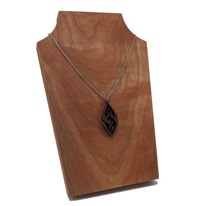 Wooden necklace pendant carved so that curving shapes weave into each other. Made from dark walnut wood and hanging from a silver chain against a cherry wood display.