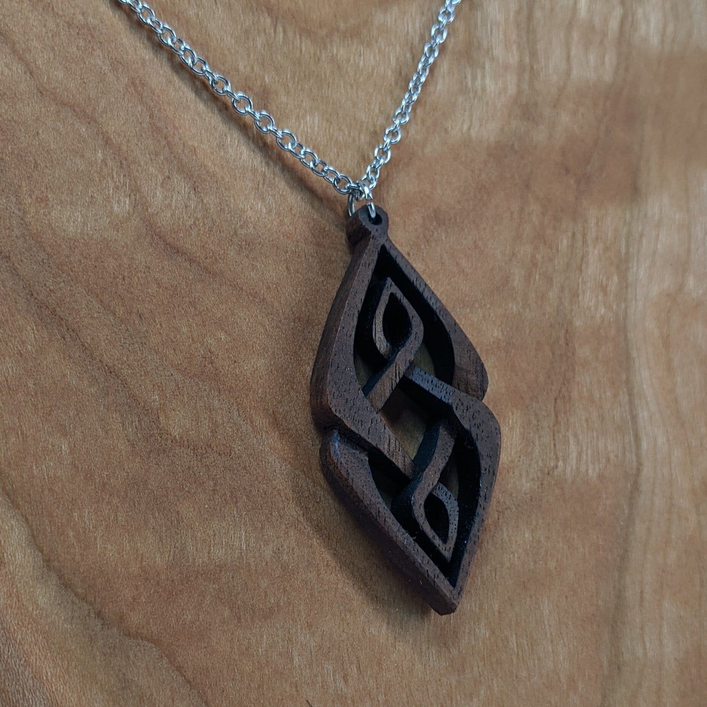 Wooden necklace pendant carved so that curving shapes weave into each other. Made from dark walnut wood and hanging from a silver chain against a cherry wood background.