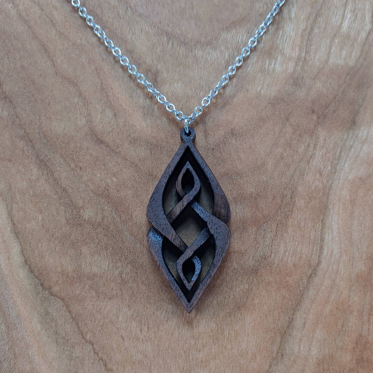 Wooden necklace pendant carved so that curving shapes weave into each other. Made from dark walnut wood and hanging from a silver chain against a cherry wood background.