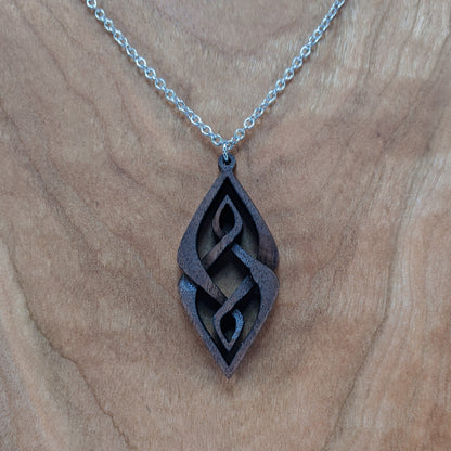 Wooden necklace pendant carved so that curving shapes weave into each other. Made from dark walnut wood and hanging from a silver chain against a cherry wood background.