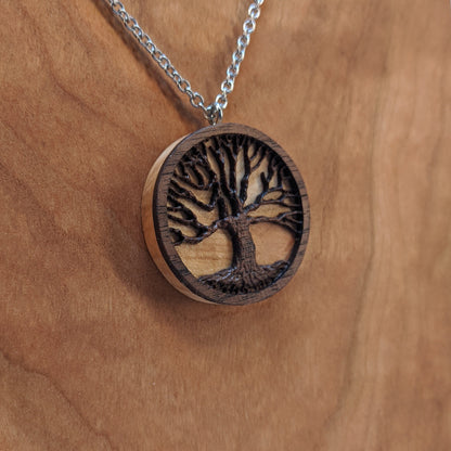 Round necklace pendant carved from contrasting hardwoods. A dark walnut tree overlayed against a light maple base. Hanging from a silver stainless steel chain against a cherry wood background.