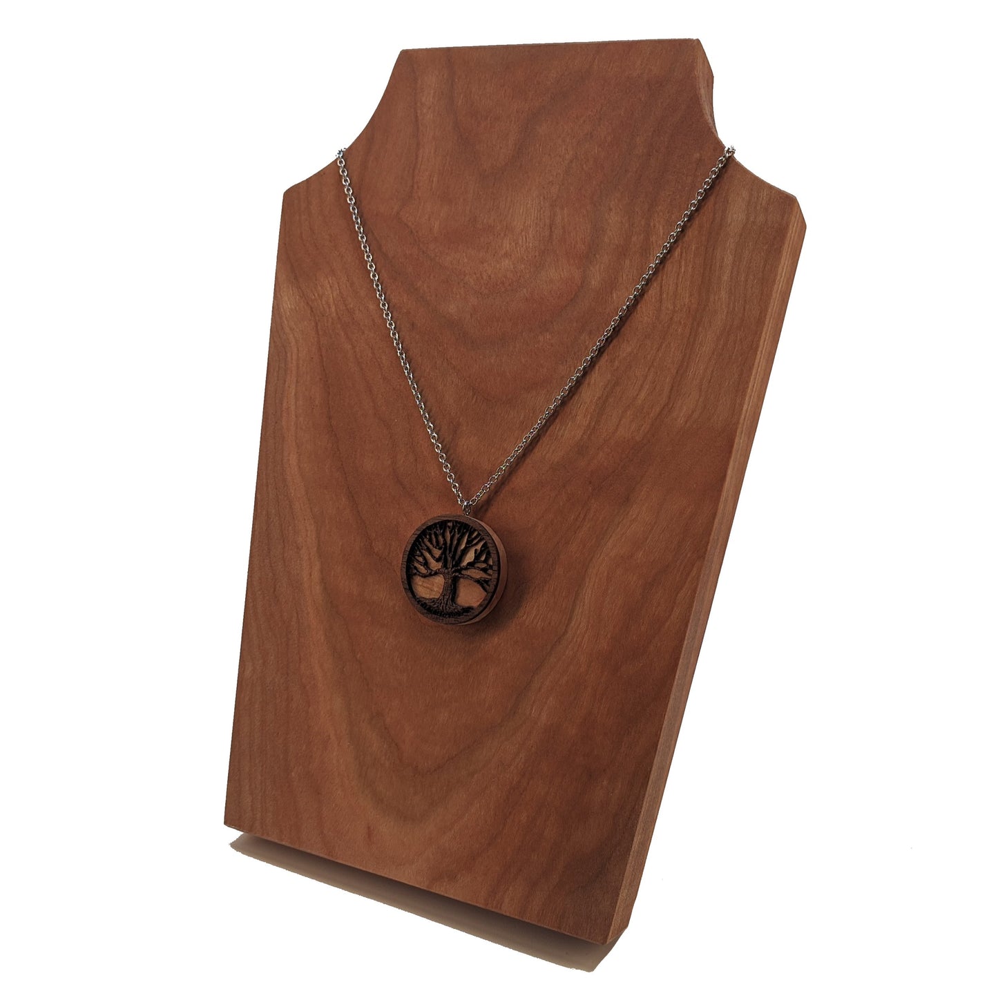 Round necklace pendant carved from contrasting hardwoods. A dark walnut tree overlayed against a light maple base. Hanging from a silver stainless steel chain against a cherry wood display.