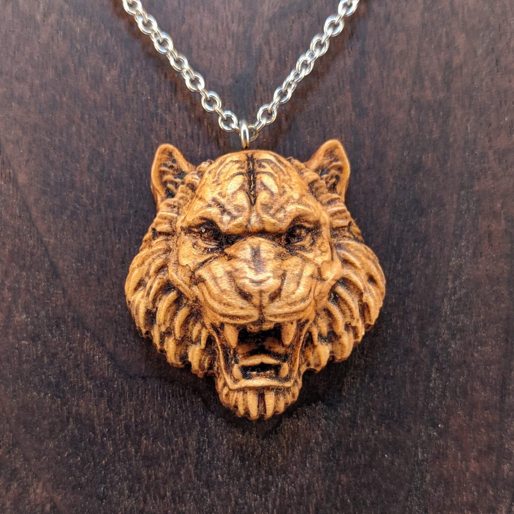 Wooden pendant necklace carved in the shape of tigers head. Made from hard maple and hanging from a black stainless steel chain against a cherry wood background.