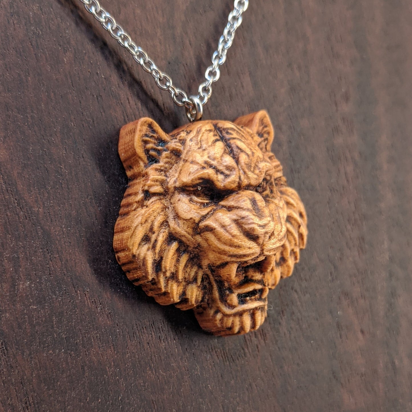 Wooden pendant necklace carved in the shape of tigers head. Made from hard maple and hanging from a silver stainless steel chain against a dark walnut background.