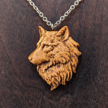 Wooden pendant necklace carved in the shape of a wolf head. The wolf faced to the side and made from hard maple wood. It hangs from a silver stainless steel chain against a dark walnut background.