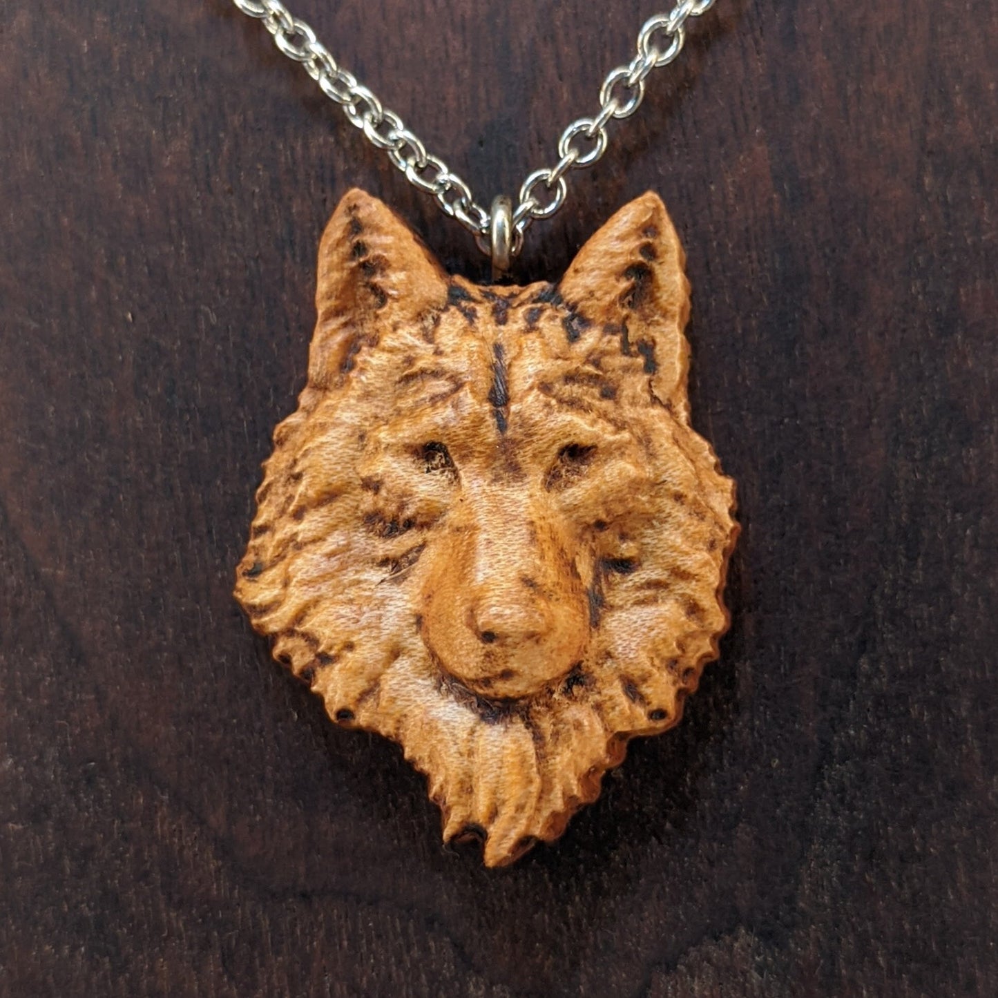 Wooden necklace pendant carved in the shaped of an wolf head. Made from hard maple wood. Hanging from a silver stainless steel chain against a dark walnut background.