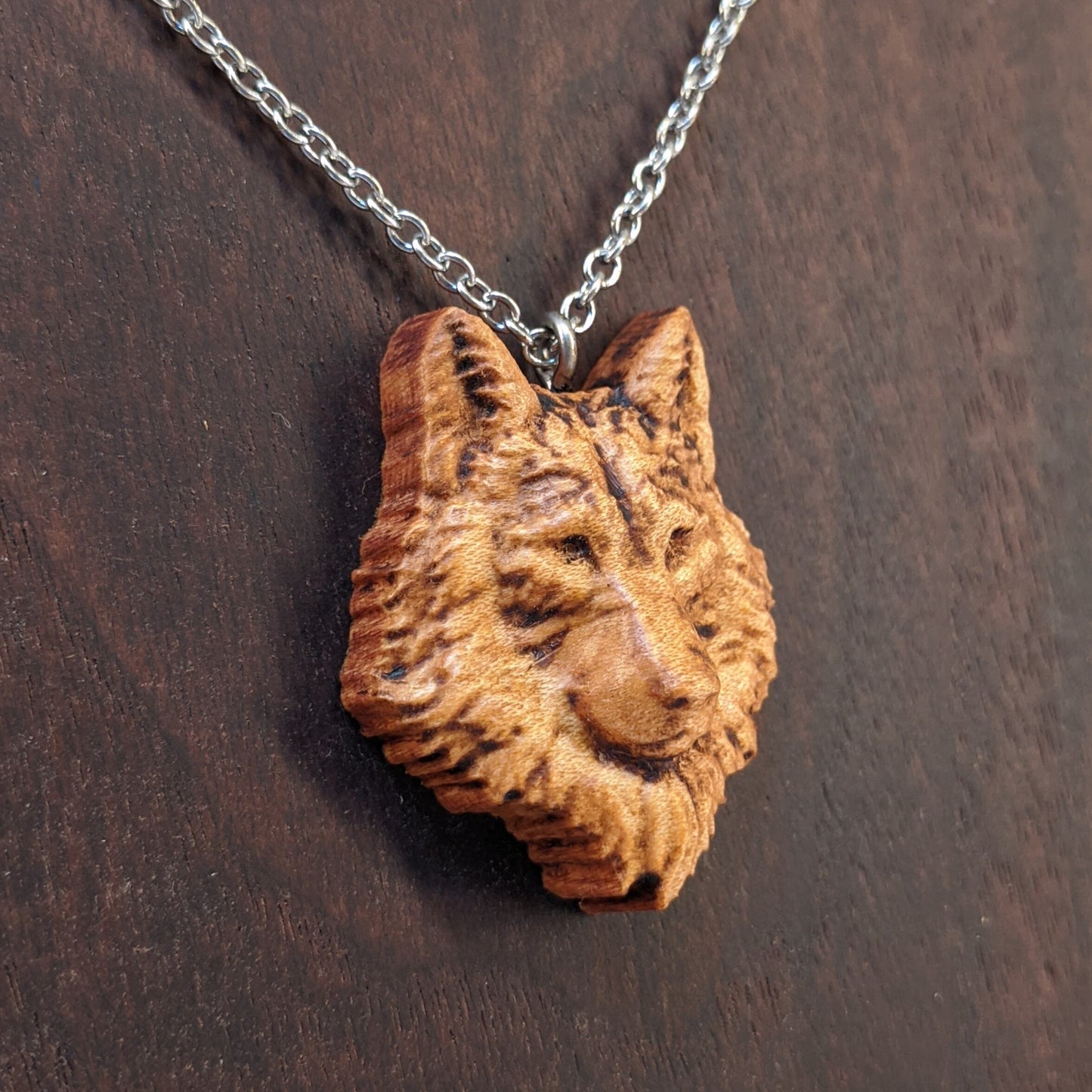 Wooden necklace pendant carved in the shaped of an wolf head. Made from hard maple wood. Hanging from a silver stainless steel chain against a dark walnut background.