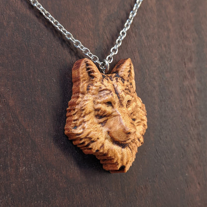 Wooden necklace pendant carved in the shaped of an wolf head. Made from hard maple wood. Hanging from a silver stainless steel chain against a dark walnut background.