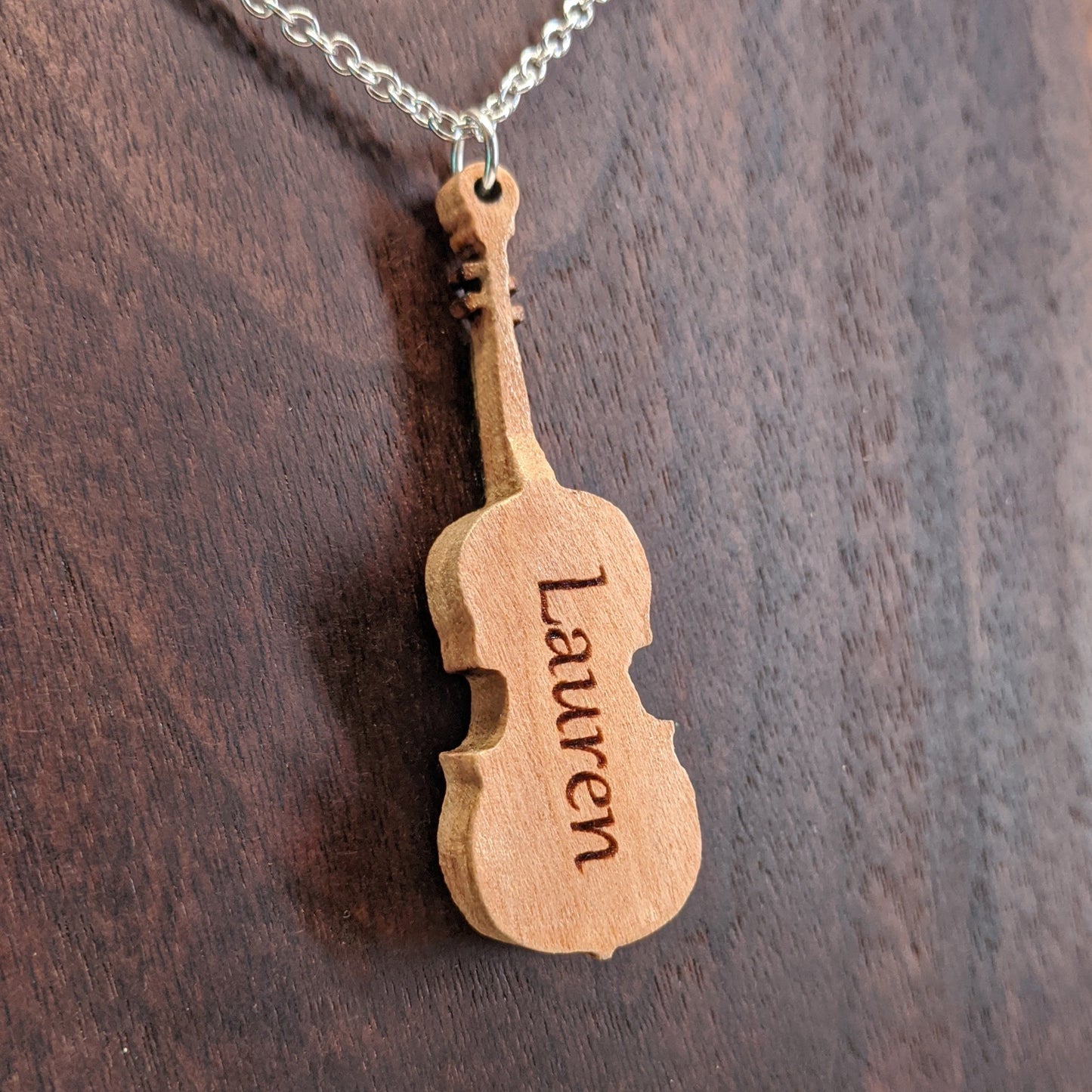 Wooden necklace pendant created in the shape of a miniature violin. The back is engraved with personalized wording. Made from hard maple wood and hanging from a silver stainless steel chain against a dark walnut background.