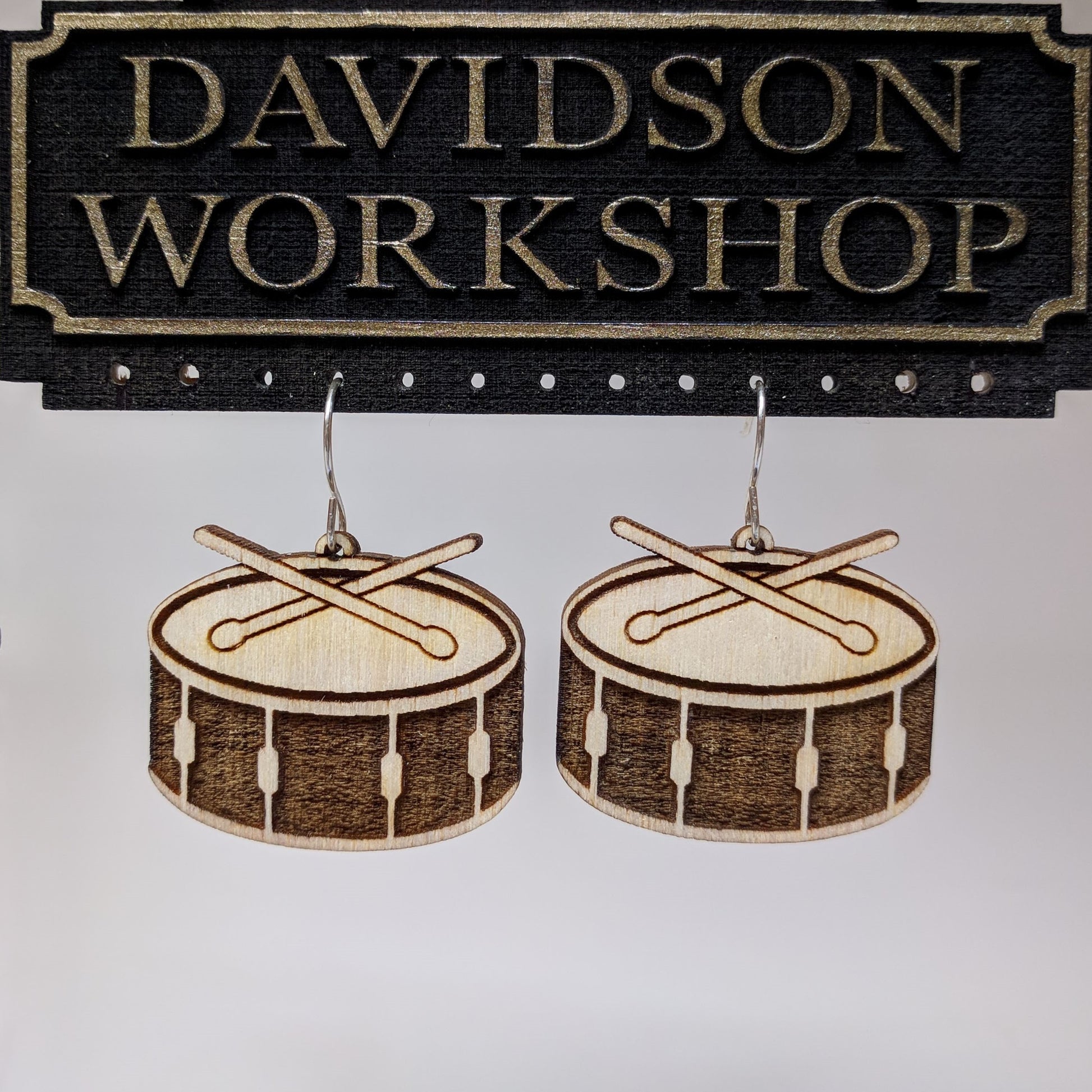 Pair of wooden earrings with silver stainless steel hooks. They are two toned light and dark brown miniature snare drums with drumsticks on top.Made from birch wood hanging from a model Davidson Workshop sign against a white background(Close up View)