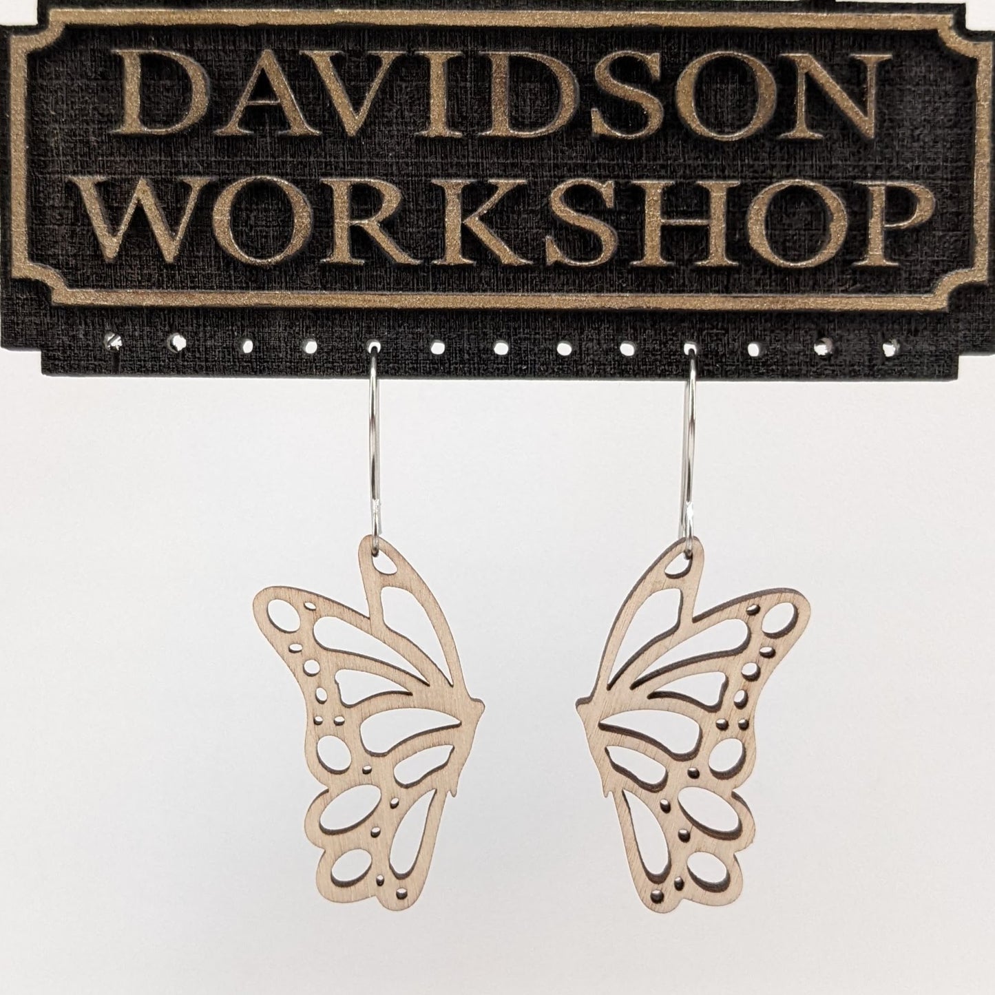 Pair of wooden earrings with silver stainless steel hooks. They are pale natural finished cutouts of side profile butterflies. Made from birch wood hanging from a model Davidson Workshop sign against a white background. (Close up View)