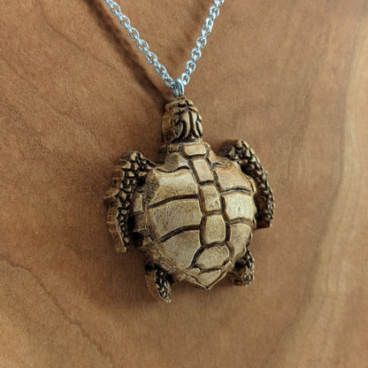 Wooden necklace pendant carved in the shape of a sea turtle. Made from hard maple wood. Hanging from a silver stainless steel chain against a cherry wood background.