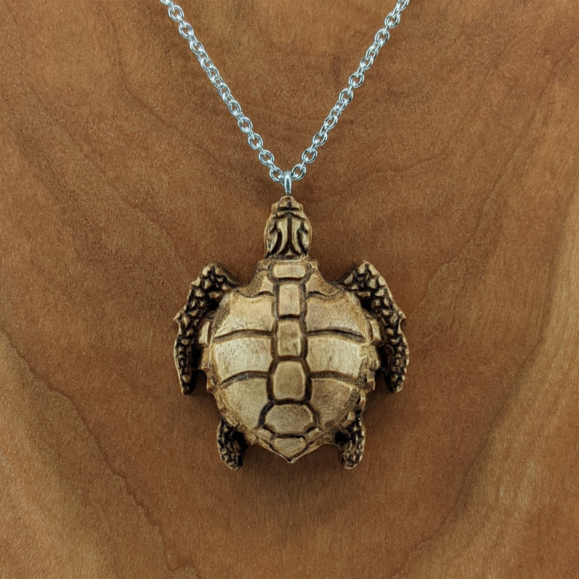 Wooden necklace pendant carved in the shape of a sea turtle. Made from hard maple wood. Hanging from a silver stainless steel chain against a cherry wood background.