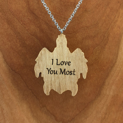 Wooden necklace pendant carved in the shape of a sea turtle. Made from hard maple wood. Hanging from a silver stainless steel chain against a cherry wood background. Showing the back, engraved with personalized wording.