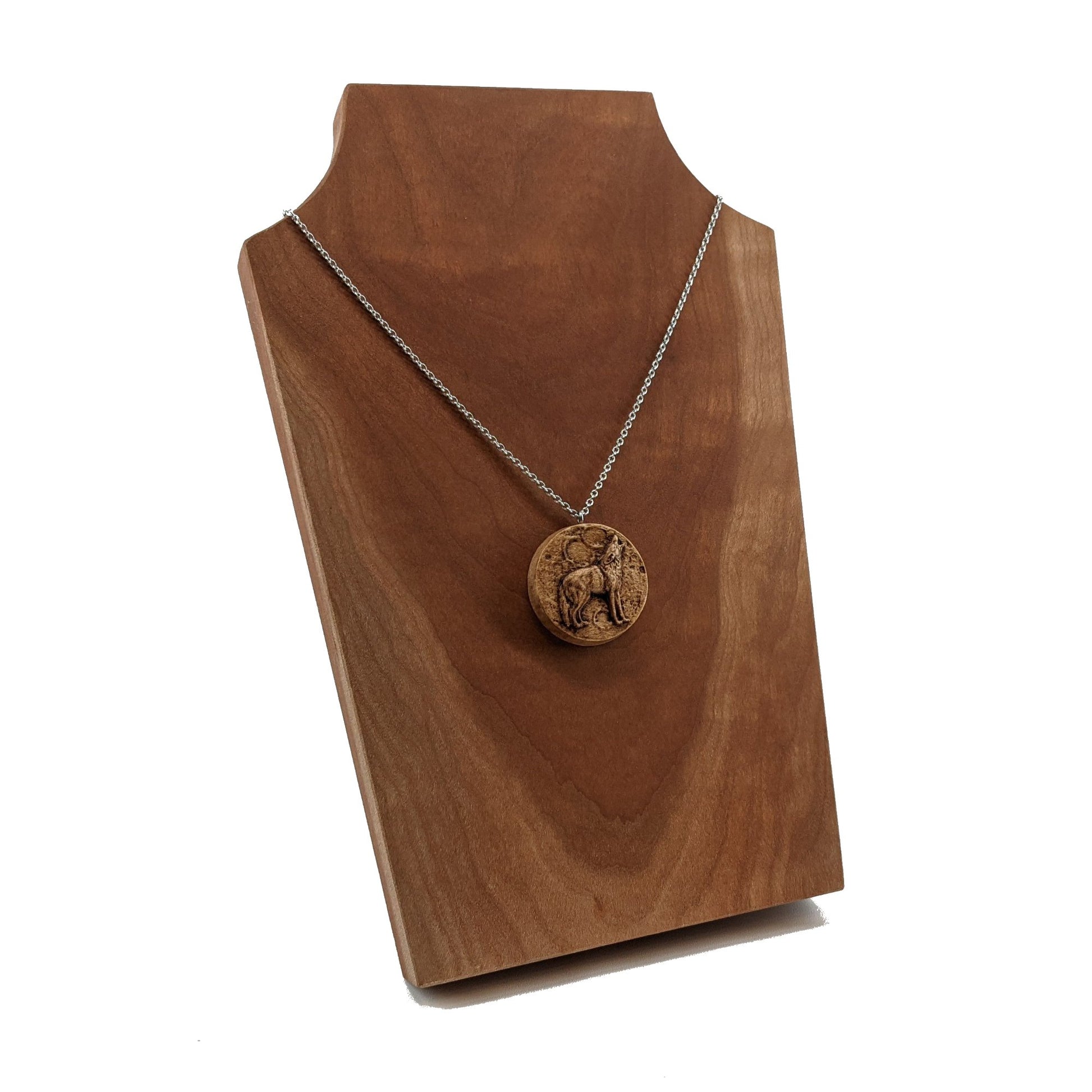 Wooden necklace pendant depicting a howling wolf in front of a full moon. Carved in a realistic style from hard maple wood. Hanging from a silver stainless steel chain against a cherry wood background.