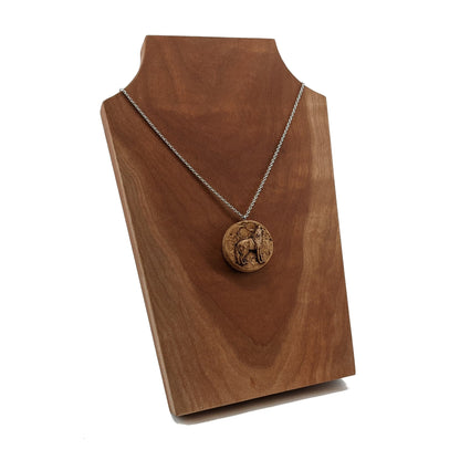 Wooden necklace pendant depicting a howling wolf in front of a full moon. Carved in a realistic style from hard maple wood. Hanging from a silver stainless steel chain against a cherry wood background.