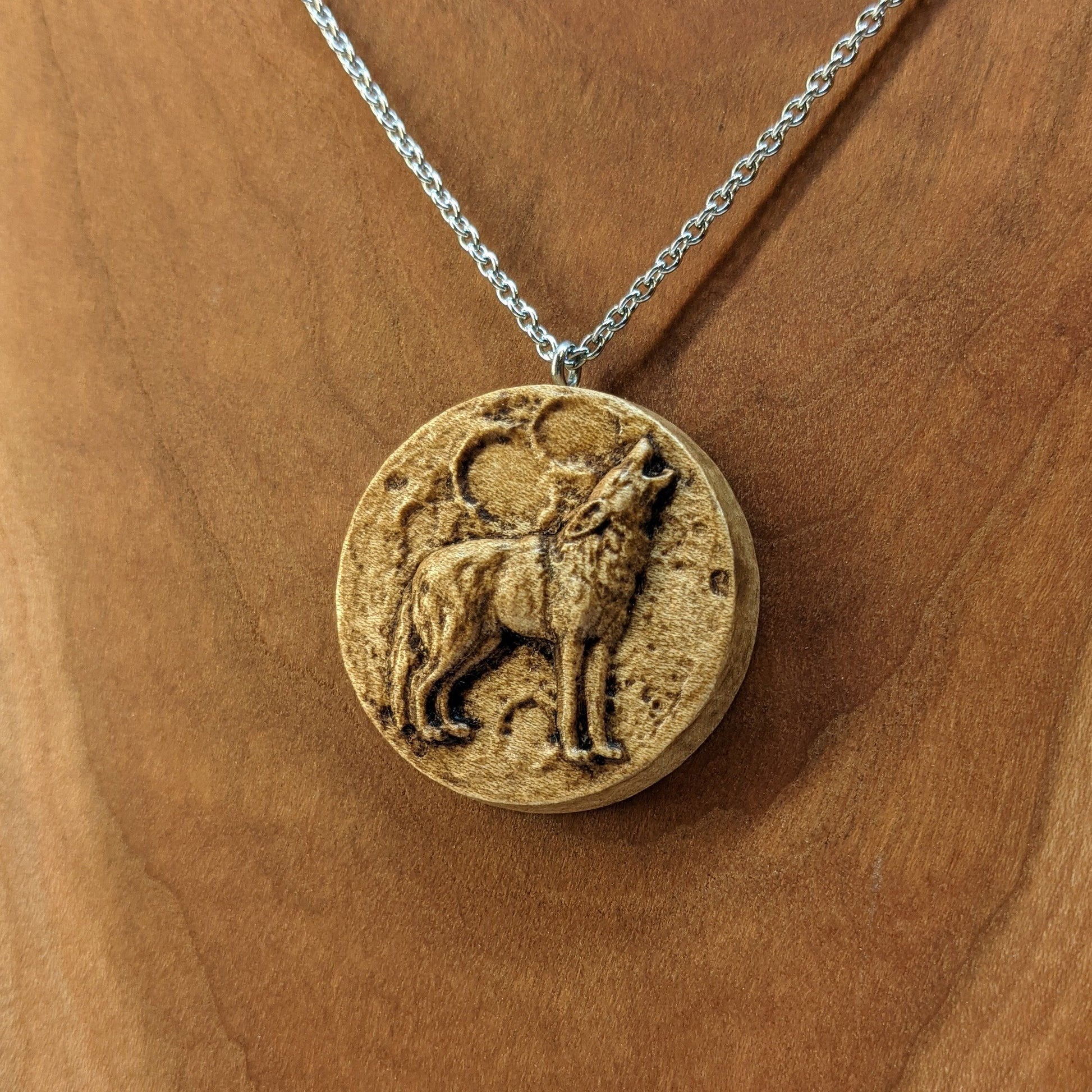 Wooden necklace pendant depicting a howling wolf in front of a full moon. Carved in a realistic style from hard maple wood. Hanging from a silver stainless steel chain against a cherry wood background.