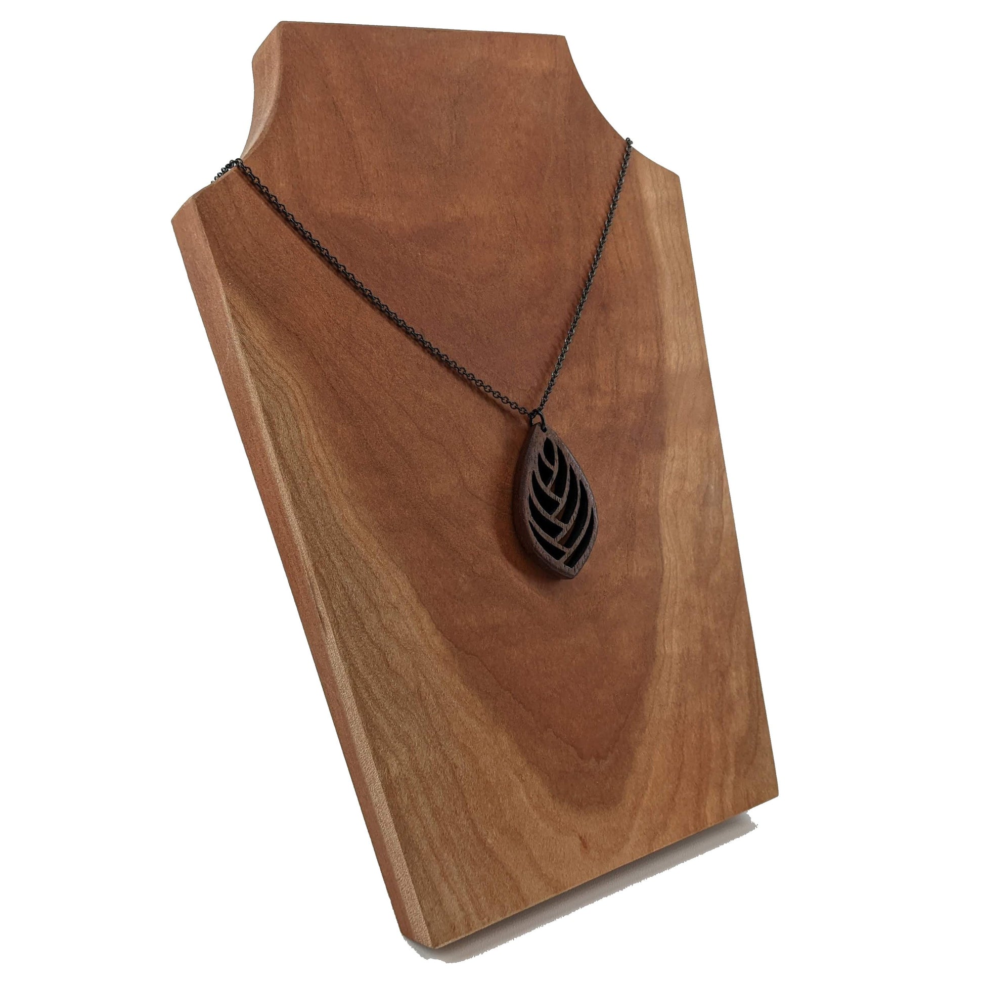 Wooden necklace pendant cut into a teardrop shape with a herring bone pattern cut throughout it. Made from walnut and hanging from a black stainless steel chain against a cherry wood background.