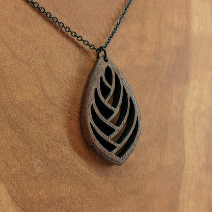 Wooden necklace pendant cut into a teardrop shape with a herring bone pattern cut throughout it. Made from walnut and hanging from a black stainless steel chain against a cherry wood background.