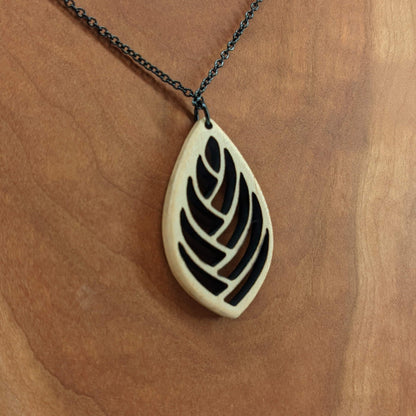 Wooden necklace pendant cut into a teardrop shape with a herring bone pattern cut throughout it. Made from hard maple and hanging from a black stainless steel chain against a cherry wood background.