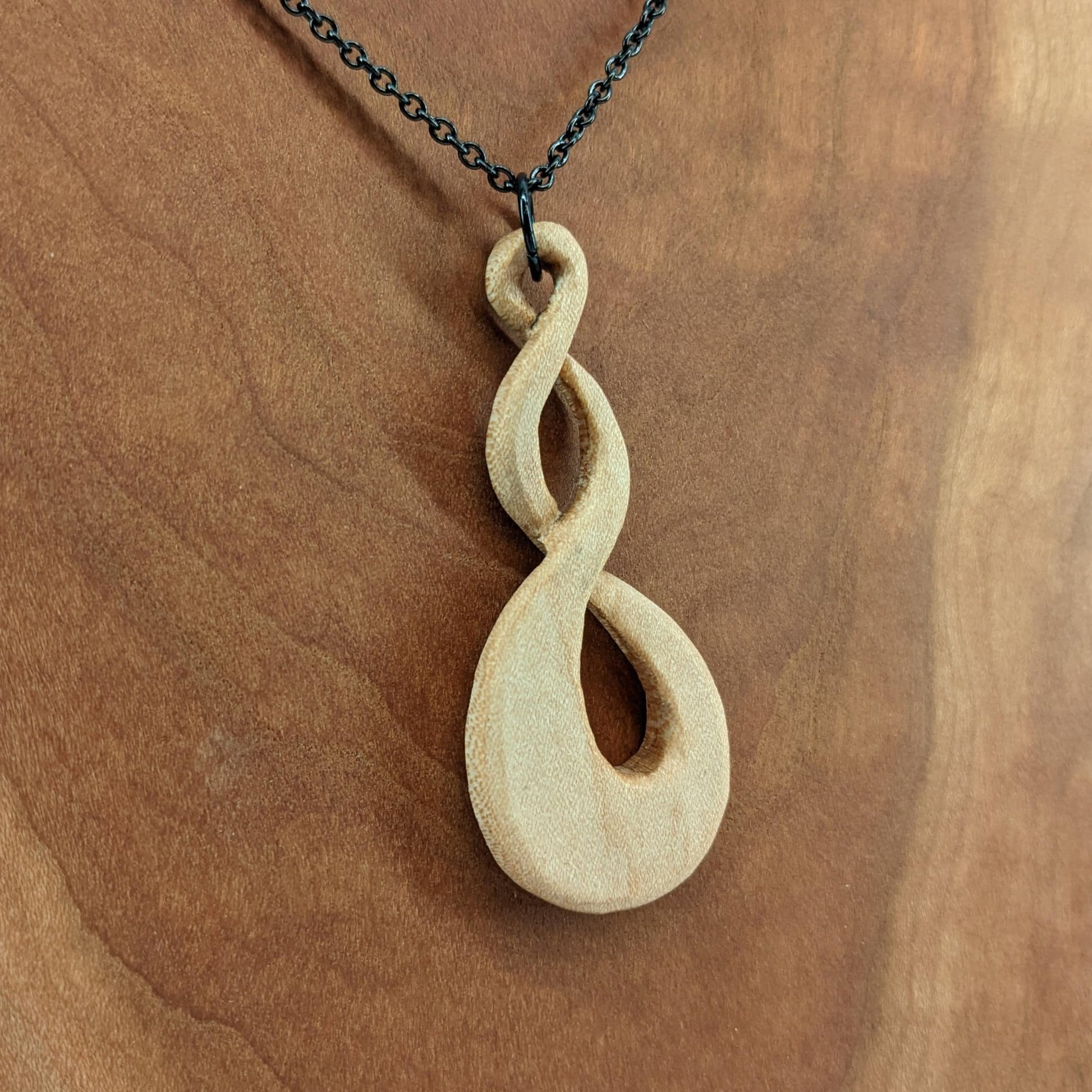 Wooden necklace pendant carved in the shape of a twisted infinity inspired symbol. Made from hard maple wood. Hanging from a black stainless steel chain against a cherry wood background.