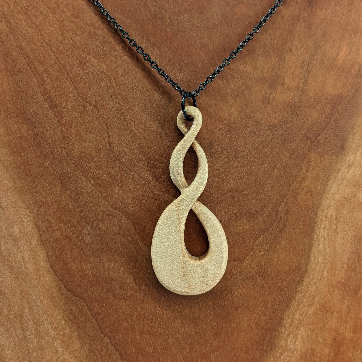 Wooden necklace pendant carved in the shape of a twisted infinity inspired symbol. Made from hard maple wood. Hanging from a black stainless steel chain against a cherry wood background.