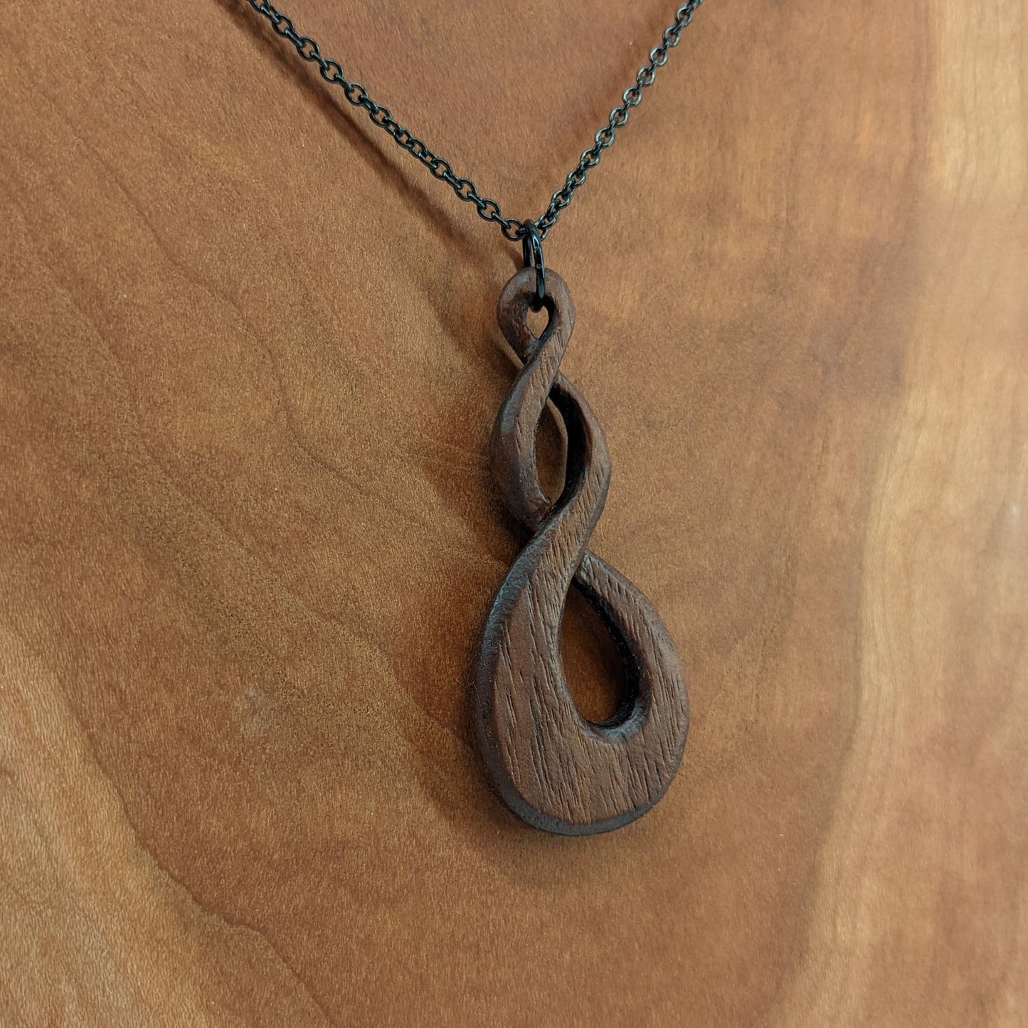 Wooden necklace pendant carved in the shape of a twisted infinity inspired symbol. Made from dark walnut wood. Hanging from a black stainless steel chain against a cherry wood background.