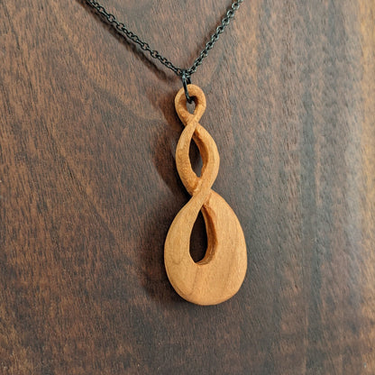 Wooden necklace pendant carved in the shape of a twisted infinity inspired symbol. Made from cherry wood. Hanging from a black stainless steel chain against a dark walnut wood background.