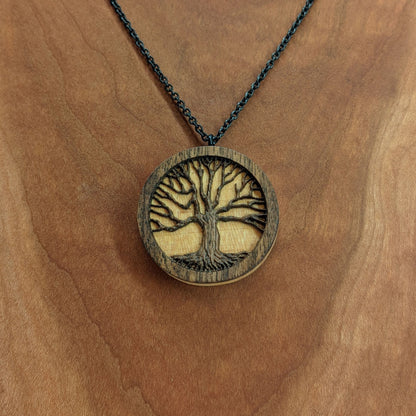 Round necklace pendant carved from contrasting hardwoods. A dark walnut tree overlayed against a light maple base. Hanging from a black stainless steel chain against a cherry wood background.