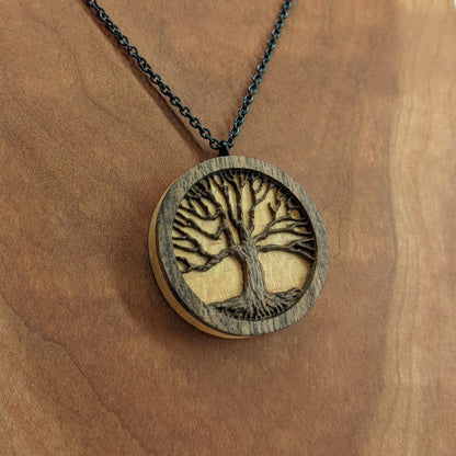 Round necklace pendant carved from contrasting hardwoods. A dark walnut tree overlayed against a light maple base. Hanging from a black stainless steel chain against a cherry wood background.