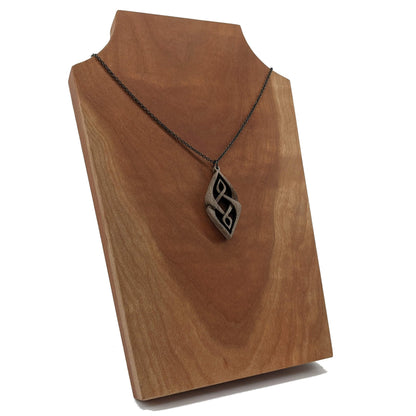 Wooden necklace pendant carved so that curving shapes weave into each other. Made from dark walnut wood and hanging from a black chain against a cherry wood display..