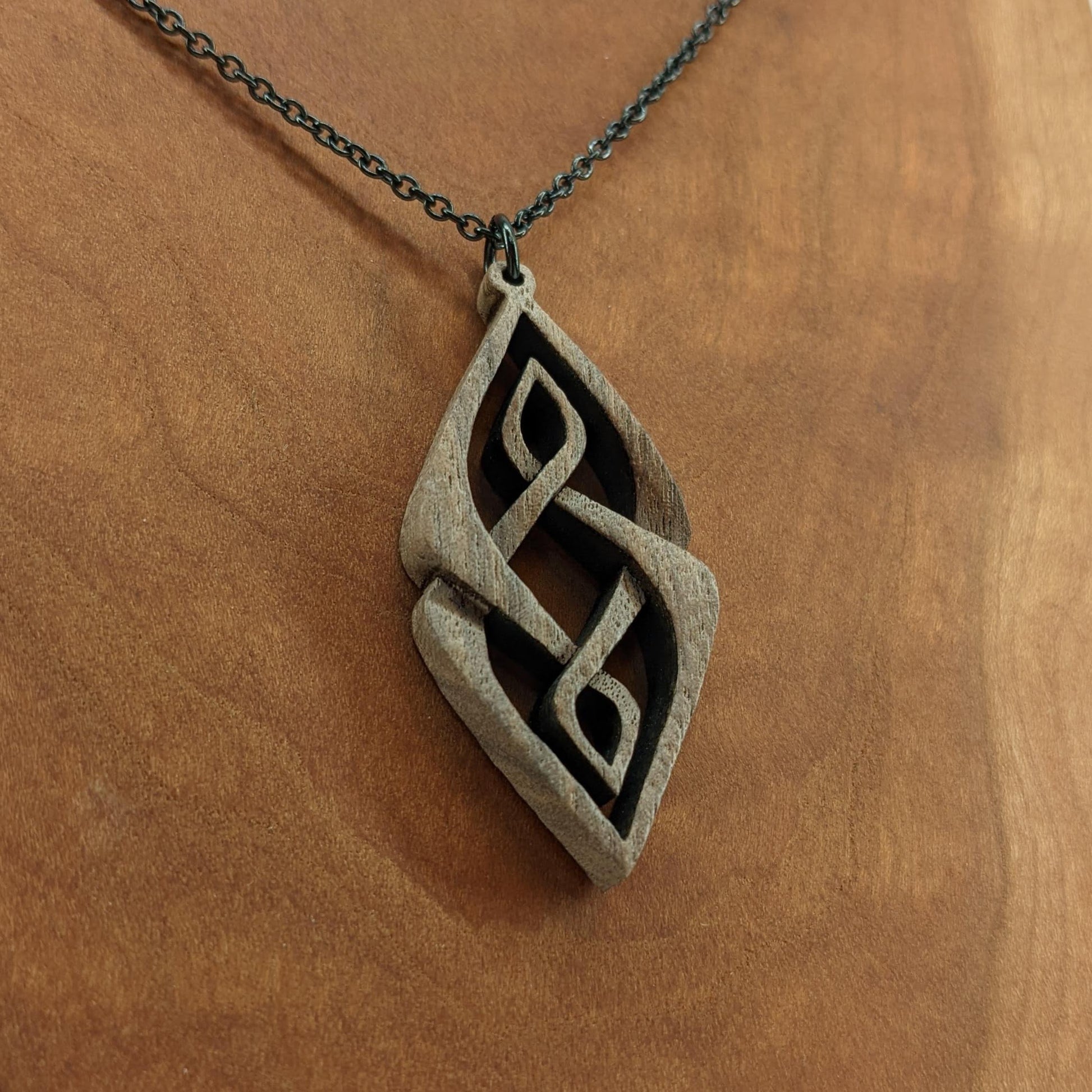 Wooden necklace pendant carved so that curving shapes weave into each other. Made from dark walnut wood and hanging from a black chain against a cherry wood background.