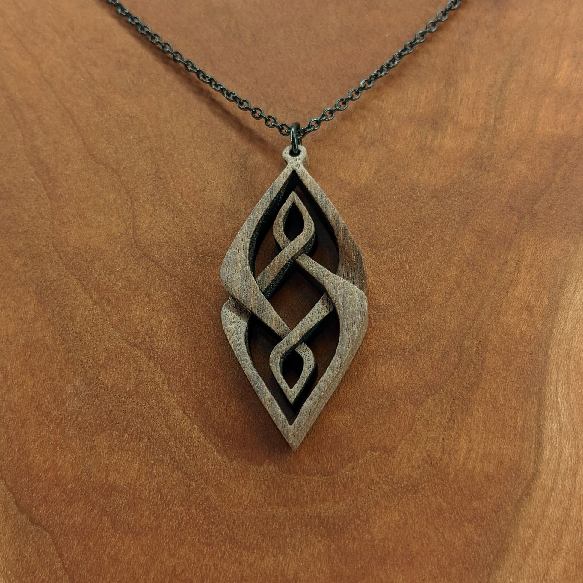 Wooden necklace pendant carved so that curving shapes weave into each other. Made from dark walnut wood and hanging from a black chain against a cherry wood background.