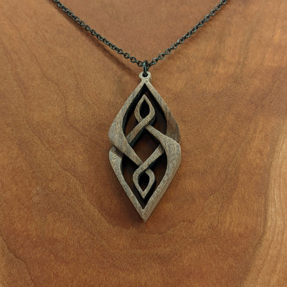 Wooden necklace pendant carved so that curving shapes weave into each other. Made from dark walnut wood and hanging from a black chain against a cherry wood background.