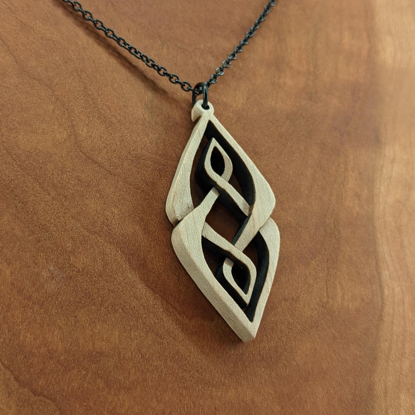 Wooden necklace pendant carved so that curving shapes weave into each other. Made from hard maple wood and hanging from a black chain against a cherry wood background.