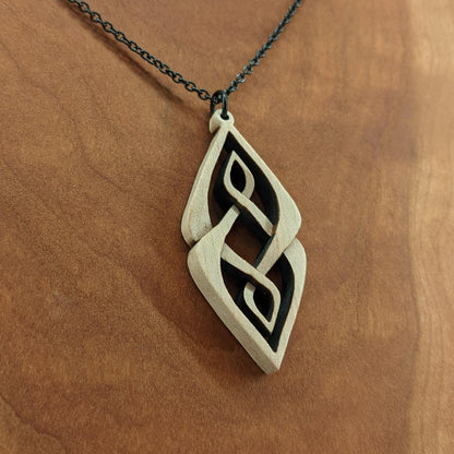 Wooden necklace pendant carved so that curving shapes weave into each other. Made from hard maple wood and hanging from a black chain against a cherry wood background.