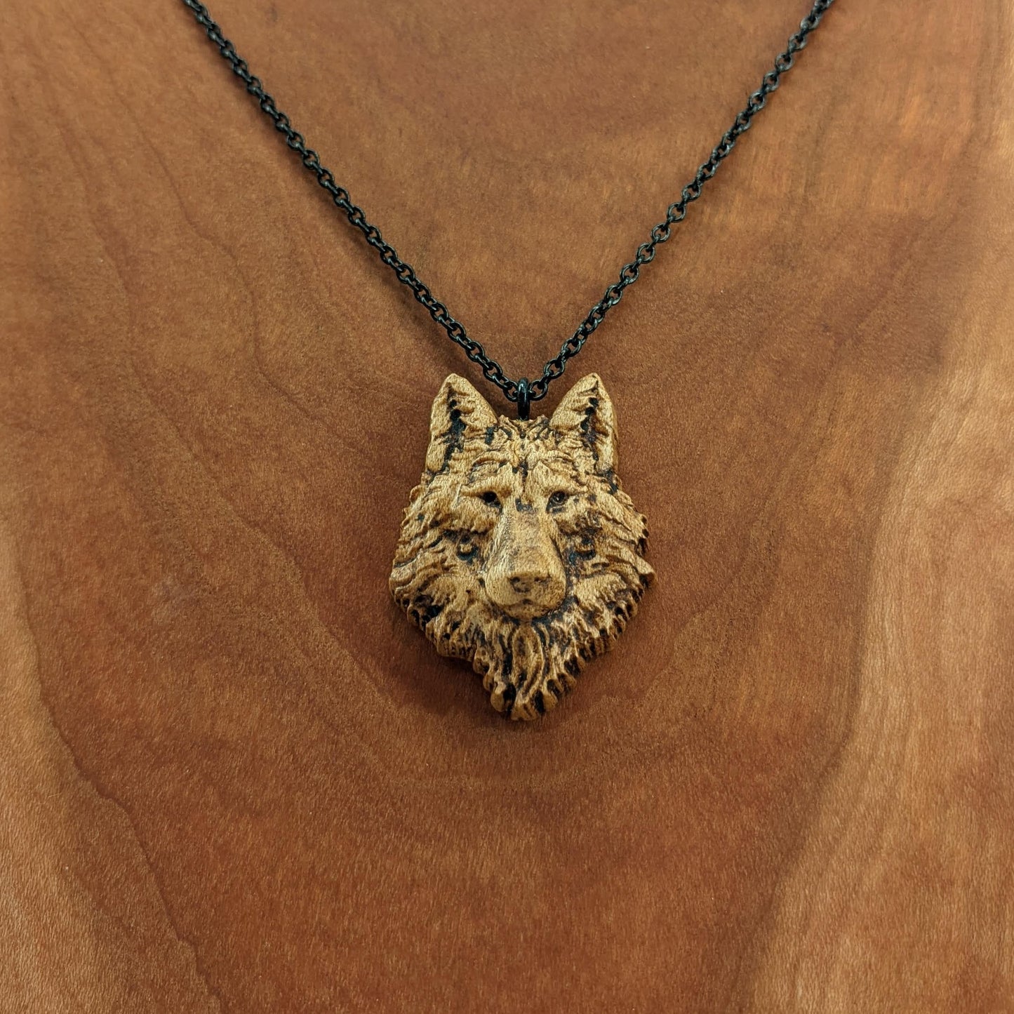 Wooden necklace pendant carved in the shaped of an wolf head. Made from hard maple wood. Hanging from a black stainless steel chain against a cherry wood background.