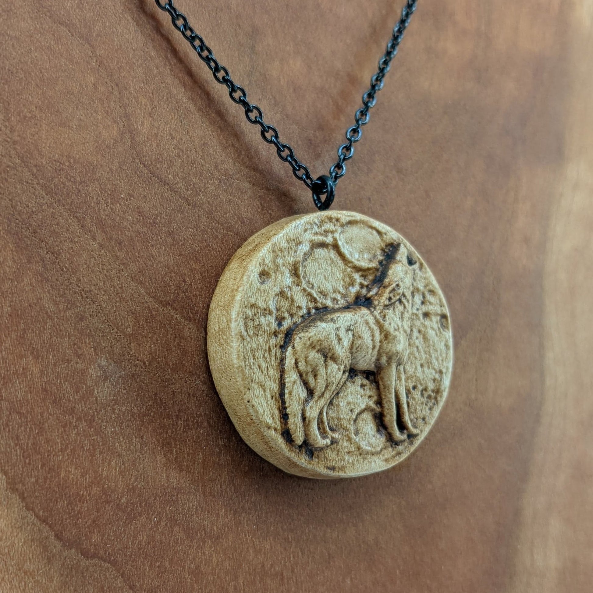 Wooden necklace pendant depicting a howling wolf in front of a full moon. Carved in a realistic style from hard maple wood. Hanging from a black stainless steel chain against a cherry wood background.