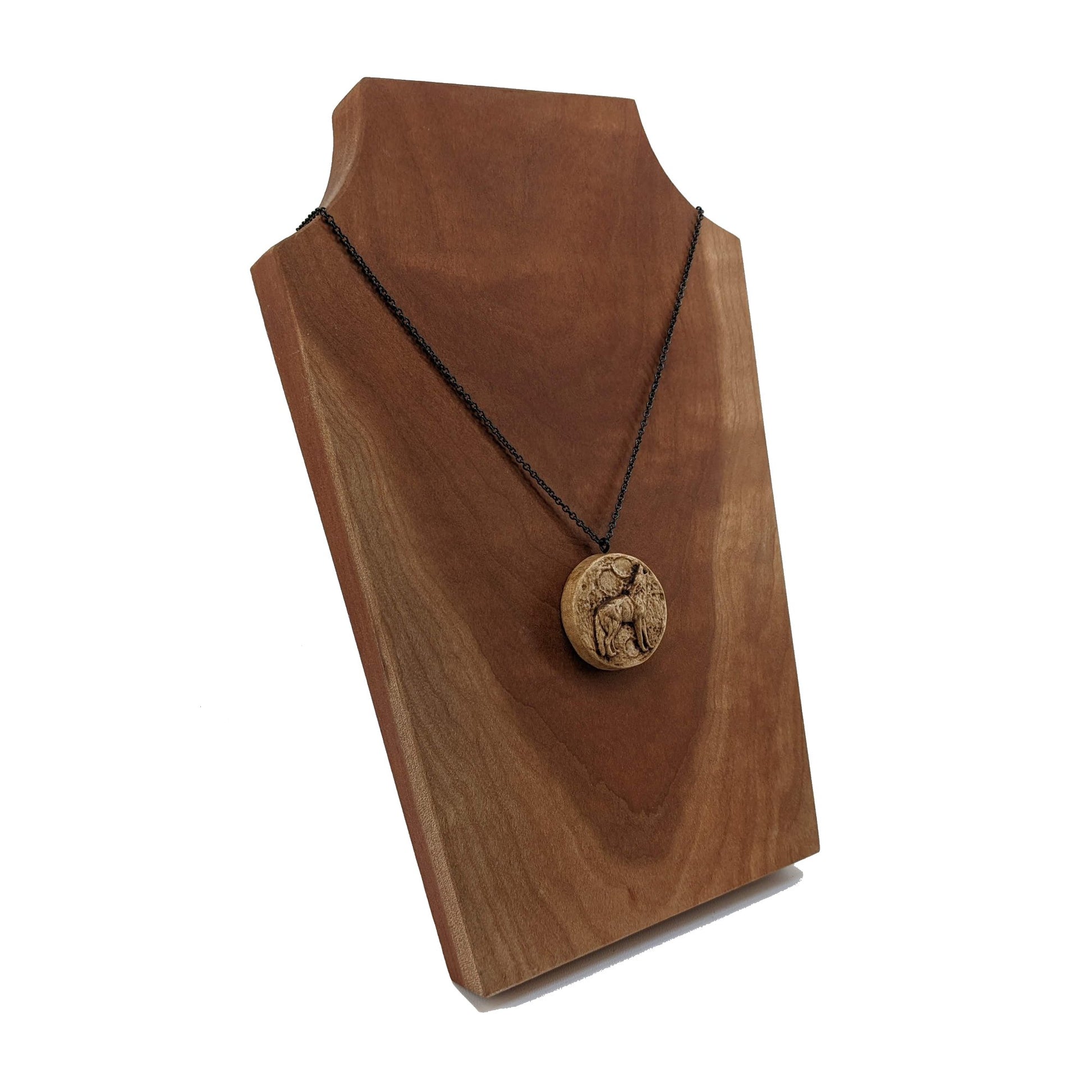 Wooden necklace pendant depicting a howling wolf in front of a full moon. Carved in a realistic style from hard maple wood. Hanging from a black stainless steel chain against a cherry wood background.