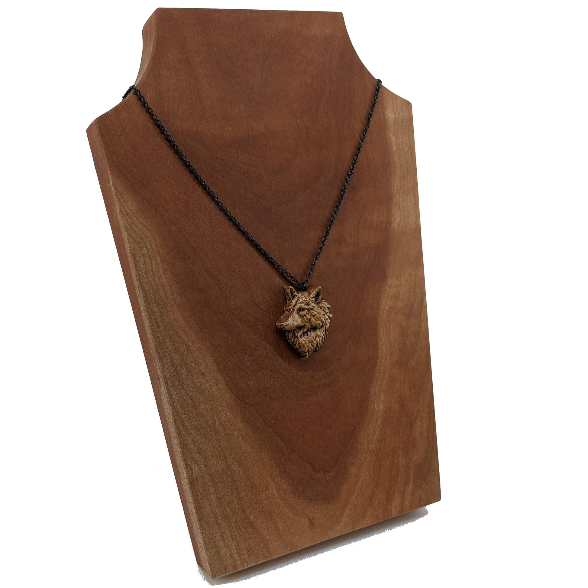 Wooden pendant necklace carved in the shape of a wolf head. The wolf faced to the side and made from hard maple wood. It hangs from a black stainless steel chain against a cherry wood background.