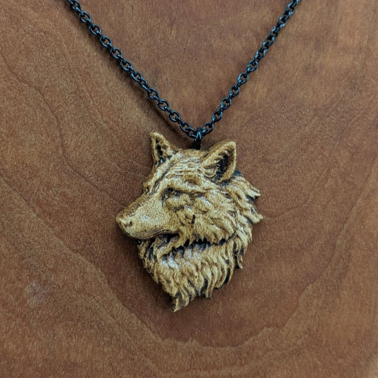 Wooden pendant necklace carved in the shape of a wolf head. The wolf faced to the side and made from hard maple wood. It hangs from a black stainless steel chain against a cherry wood background.