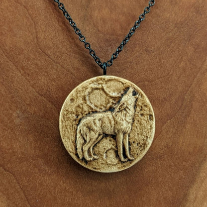 Wooden necklace pendant depicting a howling wolf in front of a full moon. Carved in a realistic style from hard maple wood. Hanging from a black stainless steel chain against a cherry wood background.