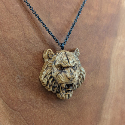 Wooden pendant necklace carved in the shape of tigers head. Made from hard maple and hanging from a black stainless steel chain against a cherry wood background.