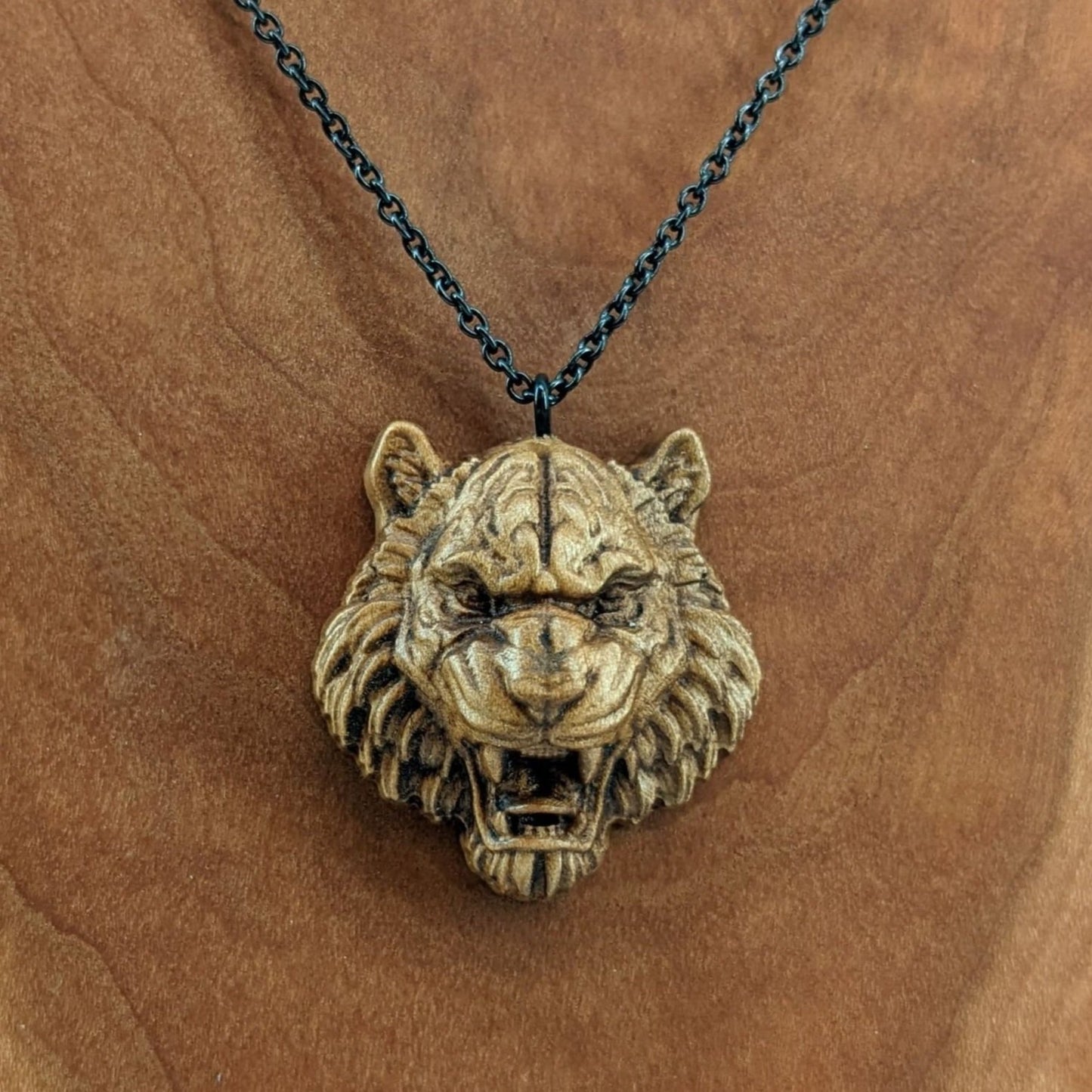 Wooden pendant necklace carved in the shape of tigers head. Made from hard maple and hanging from a black stainless steel chain against a cherry wood background.