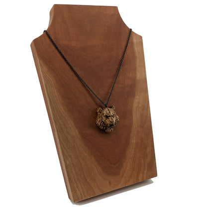 Wooden pendant necklace carved in the shape of tigers head. Made from hard maple and hanging from a black stainless steel chain against a cherry wood background.