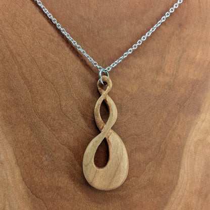 Wooden necklace pendant carved in the shape of a twisted infinity inspired symbol. Made from cherry wood. Hanging from a silver stainless steel chain against a cherry wood background.