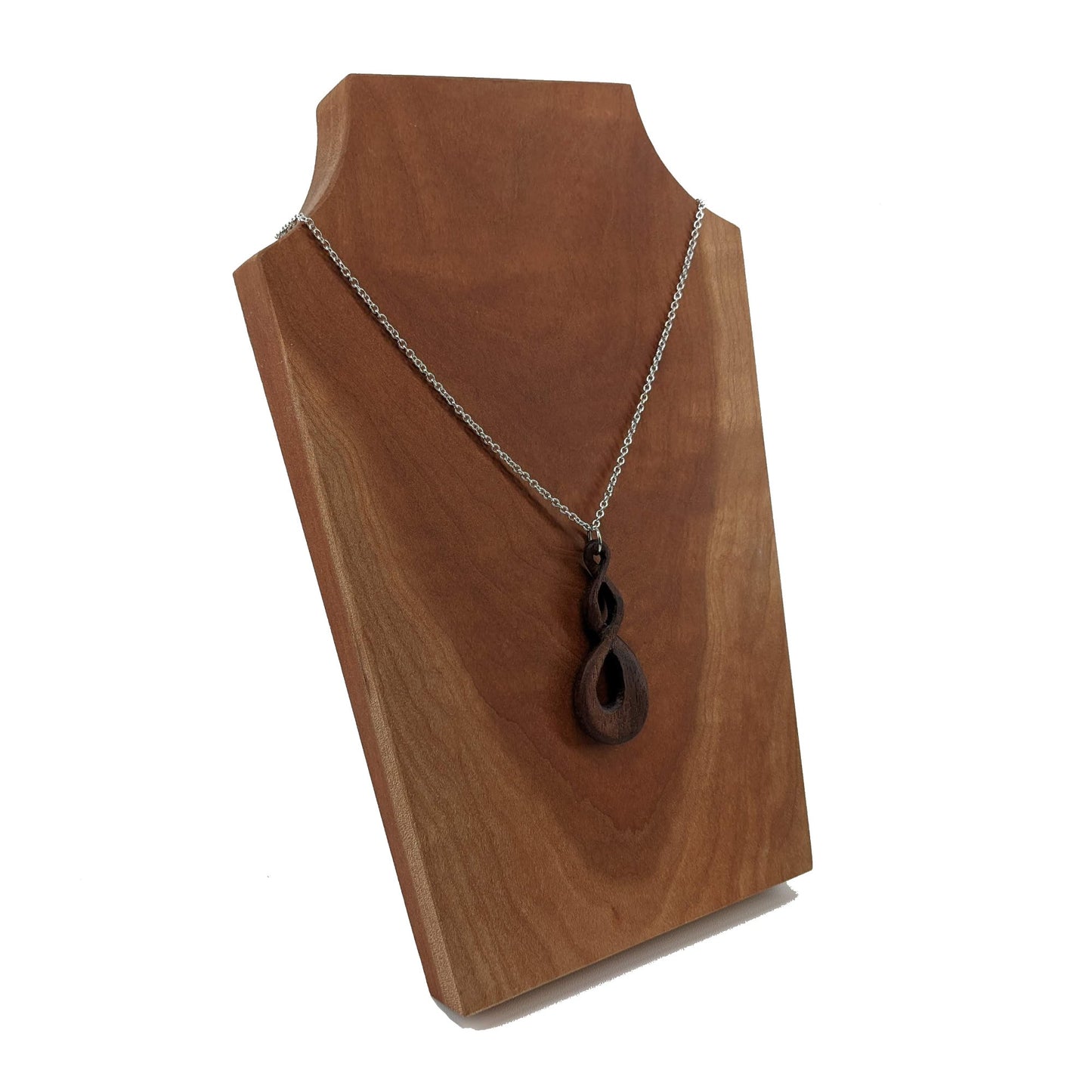 Wooden necklace pendant carved in the shape of a twisted infinity inspired symbol. Made from dark walnut wood. Hanging from a silver stainless steel chain against a cherry wood background.