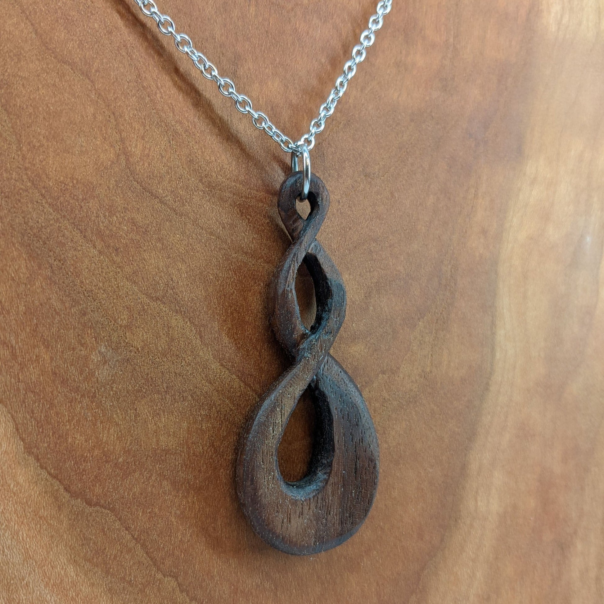 Wooden necklace pendant carved in the shape of a twisted infinity inspired symbol. Made from dark walnut wood. Hanging from a silver stainless steel chain against a cherry wood background.